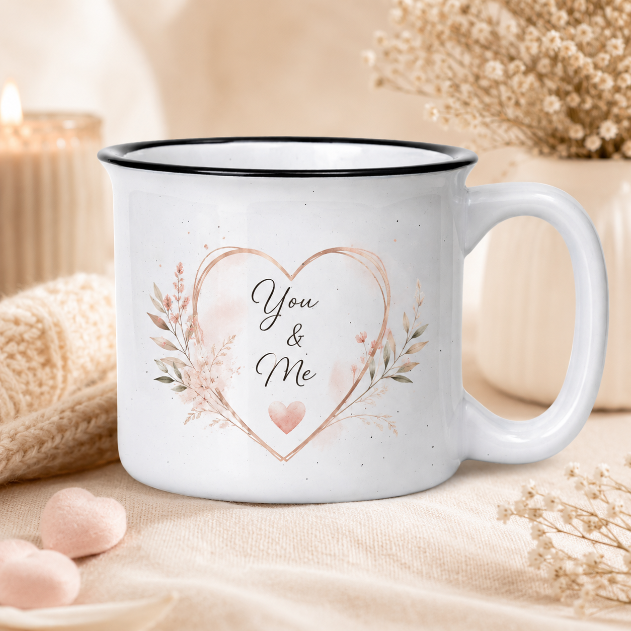 A white ceramic mug with a black rim and handle, decorated with a watercolor-style heart and floral design that reads 'You & Me'. The mug is styled on a beige fabric surface with soft decorative props in the background.